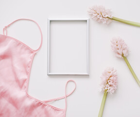 Blank photo frame, silk lingerie and pink flowers on white background. Styled feminine flat lay, top view, copy space. Creative concept, empty greeting card