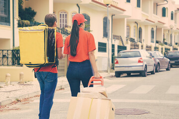 Wall Mural - Two post office workers with yellow thermal bag and boxes on trolley. Back view of couriers in red shirts looking for address and delivering order. Delivery service and online shopping concept