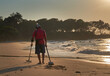 A man is searching valuables with a metal detector on a beach in Thailand during sunset.