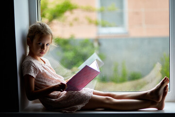 Cute little girl reading book at home, at windowsill