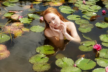 Portrait Of Sexy Young Redhead Woman Mermaid Sits Seductive With Pink Water Lilies In The Water, Lake, Pond