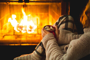 Relaxed young woman in pyjamas sitting near a chimney fire enjoying a warm cup of hot chocolate. Concept about lifestyle and people.