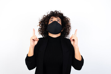 Wall Mural - young beautiful caucasian woman wearing medical mask standing against white wall being amazed and surprised looking and pointing up with fingers showing something strange.