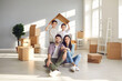 © Studio Romantic - Portrait of happy young family in new home. Smiling mom and dad sitting on floor with unpacked boxes, and children holding symbolic roof behind. Real estate, mortgage, buying and moving house concept