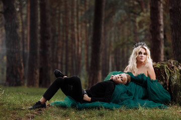 A beautiful blonde young woman in a long green dress and a diadem on her head with stylish young son in the forest. girl and boy sitting near the old stump. Solar glare. Fantasy. fairy tale