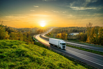 Wall Mural - White truck driving on the highway winding through forested landscape in autumn colors at sunset