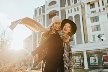 Wall Mural - Pretty girl in long scarf enjoying weekend with friend. Outdoor photo of two joyful ladies fooling around in autumn.