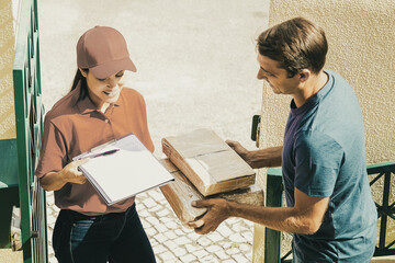 Wall Mural - Cheerful post office worker delivering carton boxes to client, smiling and holding clipboard for signing. Attractive man receiving order at home. Express delivery service and online shopping concept