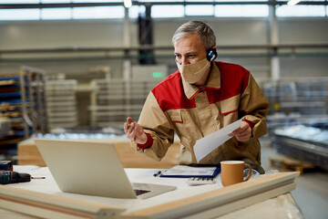 Wall Mural - Happy worker with face mask talking during video call over laptop at carpentry workshop.