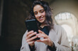 © Roman - Low angle shot of young good-looking smiling woman wearing mask and light-colored coat. Good vibes while walking city streets on sunny day and using smartphone to chat. Modern technology advantages