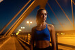 © Roman - Young female runner in blue top confidently standing on bridge at twilight. Attractive woman working out outdoors to help maintain healthy weight. Physical activity and weight management concept