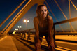 © Roman - Young female athlete stopped to regain breath after running over bridge at twilight. Sportswoman puts her hands on her knees after morning jogging routine to recover. Sports and health concept