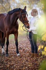 Young girl is standing with her horse on a forest path in autumn and the horse is sniffing at her sweater..