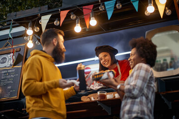 Wall Mural - female employee socializing with multiethnic couple eating sandwiches in front of fast food service, talking, smiling