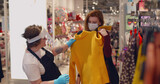 Fototapeta  - Shop assistant in safety mask and gloves showing sweater to beautiful girl in the department store