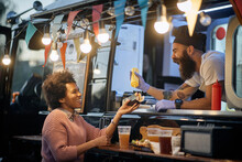 Young Beardy Caucasian Employee In Fast Food Laughing, Adding A  Mustard In A Sandwich To A Female Afro-american Customer