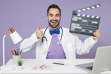 Wall Mural - Smiling male doctor man in white medical suit sits at desk in hospital clinic office hold classic black film making clapperboard show thumb up gesture isolated on violet background studio portrait.