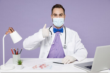 Wall Mural - Male doctor man in white medical suit sit at desk work in clinic office in sterile face mask, from coronavirus virus covid-19, latex gloves show thumb up isolated on violet background studio portrait