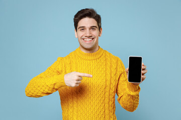 Young smiling happy student man 20s in knited yellow sweater hold in hand using point index finger on mobile cell phone with blank screen workspace area isolated on blue background studio portrait.