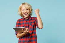Happy Little Curly Kid Boy 10s Years Old In Basic Red Checkered Shirt Using Tablet Pc Computer Doing Winner Gesture Isolated On Blue Background Children Studio Portrait. Childhood Lifestyle Concept.