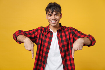 Young spanish latinos smiling handsome fashionable man 20s in red checkered shirt look camera pointing down on workspace area copy space mock up isolated on yellow color background studio portrait.