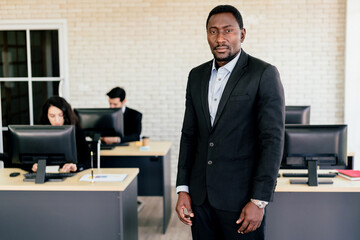 confident african american businessman standing in office with blur employees working on their desk 