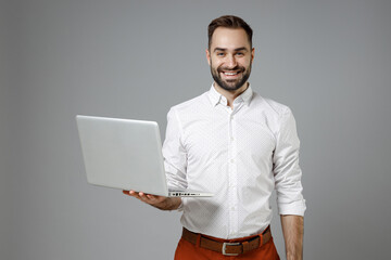 Canvas Print - Smiling handsome young bearded business man in classic white shirt working on laptop pc computer looking camera isolated on grey color background studio portrait. Achievement career wealth concept.