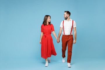 Full length smiling young couple two friends man woman in white red clothes holding hands looking at each other isolated on pastel blue background studio portrait. St. Valentine's Day holiday concept.