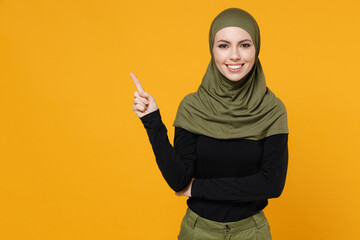 Wall Mural - Smiling young arabian muslim woman in hijab black green clothes pointing index finger up on mock up copy space isolated on yellow color background, studio portrait. People religious lifestyle concept.