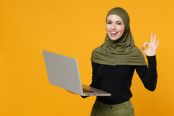 Wall Mural - Funny young arabian muslim woman in hijab black green clothes working on laptop pc computer showing OK gesture isolated on yellow color background studio portrait. People religious lifestyle concept.