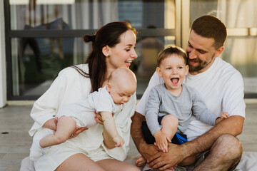 A young beautiful family with two little sons hug and kiss in the yard of their own house. Young mom and dad with two young sons hug and kiss in the yard of their own house.