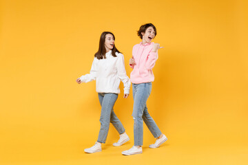 Wall Mural - Full length side view of excited shocked two young women friends 20s wearing casual white pink hoodies walking going pointing thumb aside isolated on bright yellow color background studio portrait.