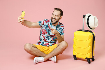 Full length laughing young traveler tourist man sit on floor doing selfie shot point index finger on mobile phone isolated on pink background. Passenger travel on weekend. Air flight journey concept.