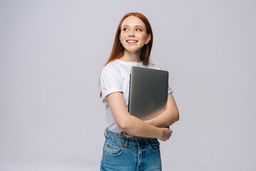 Smiling young woman student holding laptop computer and looking away on isolated gray background. Pretty lady model with red hair emotionally showing facial expressions in studio, copy space.