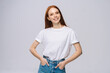 © dikushin - Cheerful young woman wearing T-shirt and denim pants holds hands in pockets on isolated white background. Pretty lady model with red hair emotionally showing facial expressions in studio, copyspace.