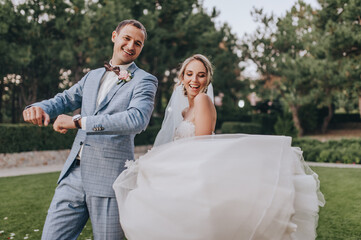 Stylish groom in a gray checkered suit and a blonde bride in a long white dress are cheerfully and funny dancing in a clearing with green grass, against the background of the forest. Wedding portrait.