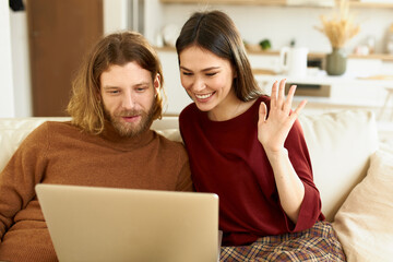 Recreation, connection and technology. Attractive unshaven young male relaxing at home with his pretty wife, seated on sofa with open laptop, waving hand and smiling, having video conference call