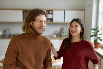 Handsome young bearded guy laughing at joke while having fun at home, speaking to his cute cheerful girlfriend, fooling around, expressing positive emotions. People, lifestyle and relationships