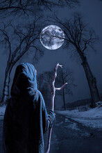 A Witch With A Stick Walks Down A Narrow Street On A Winter Night. A Big Full Moon Shines Through Tall Trees.