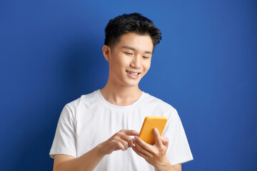 Wall Mural - Casually dressed student standing isolated on blue background looking at screen of smart phone, browsing web pages and smiling while chatting with friends