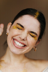 Wall Mural - Close-up portrait of gorgeous female model in golden accessories. Studio photo of attractive brunette girl laughing with eyes closed.