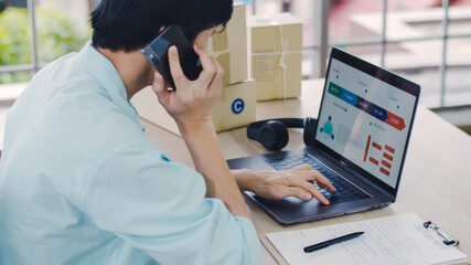 Asian man small business owner using laptop checking statistic graph top seller product at the home office and using smartphone during the covid-19 pandemic.