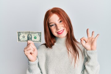 Wall Mural - Young red head girl holding 1 dollar banknote doing ok sign with fingers, smiling friendly gesturing excellent symbol