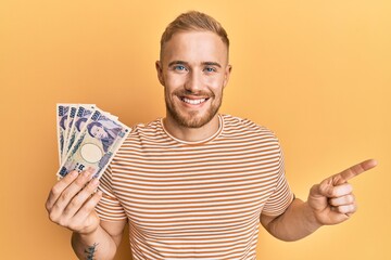 Wall Mural - Young caucasian man holding japanese yen banknotes smiling happy pointing with hand and finger to the side