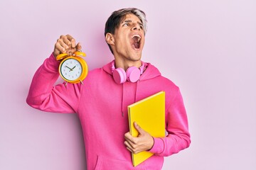 Young hispanic man holding book and alarm clock angry and mad screaming frustrated and furious, shouting with anger looking up.