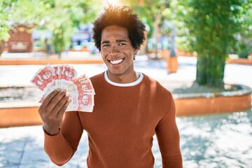 Wall Mural - Young african american man smiling happy. Standing with smile on face holding colombian pesos banknotes at town street.