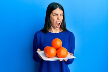 Wall Mural - Young hispanic woman holding plate with fresh oranges angry and mad screaming frustrated and furious, shouting with anger. rage and aggressive concept.