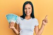Beautiful hispanic woman holding 100 brazilian real banknotes smiling happy pointing with hand and finger to the side