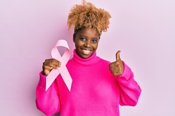 Sticker - Young african woman with afro hair holding pink cancer ribbon smiling happy and positive, thumb up doing excellent and approval sign