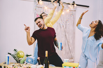 Happy hipster guy in cone gat having fun singing greeting song on birthday party at home with festive interior, group of best friends making anniversary celebration great feeling amazed together